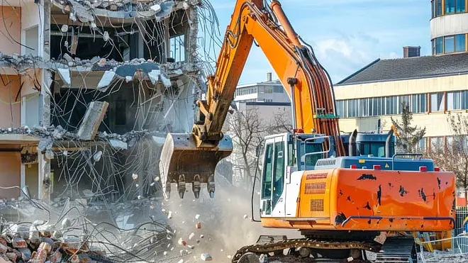Ein Kettenbagger reißt ein Gebäude ab, während Beton und Bauschutt auf einer Baustelle herunterfallen.