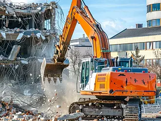Ein Kettenbagger reißt ein Gebäude ab, während Beton und Bauschutt auf einer Baustelle herunterfallen.